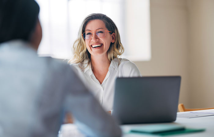 A woman smiling, sitting in front of a laptop while meeting with another person