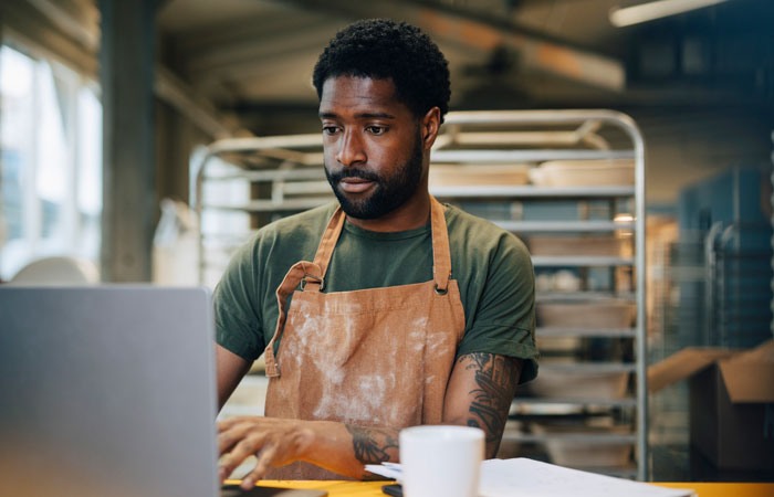 A man in a bakery sitting at a table working on a laptop