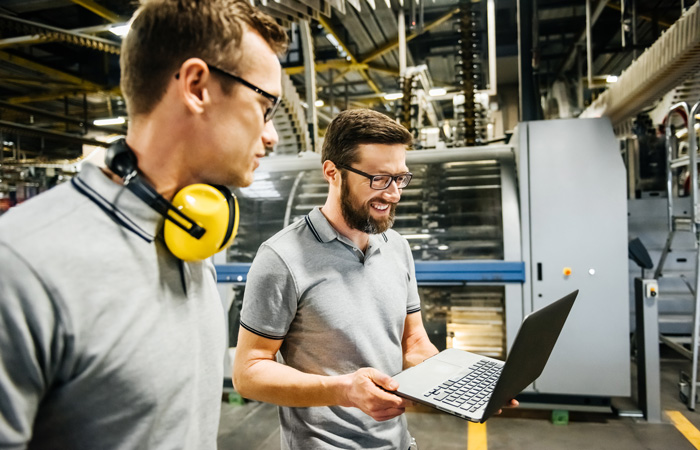 Two warehouse employees working on a laptop in front of machinery