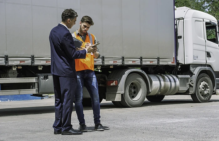 A driver and a man in a suit standing next to a semi truck looking at a clipboard.