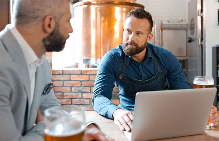 Two men in a brewery talking while working on a laptop