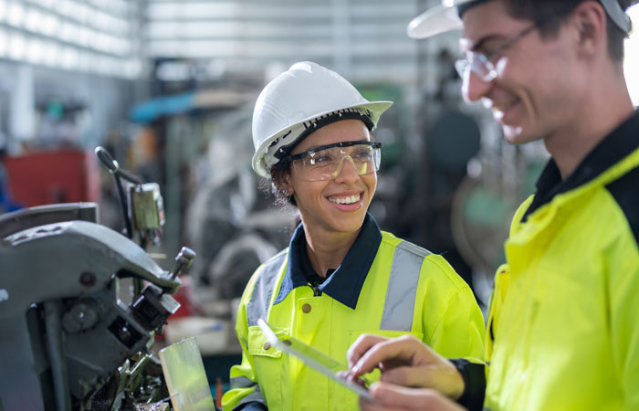 Two people smiling wearing hi-viz gear, hard hats, and eye protection while looking at machinery 