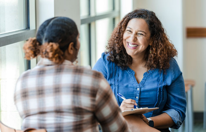 Two women sitting at a table talking. One is holding a clipboard and smiling.