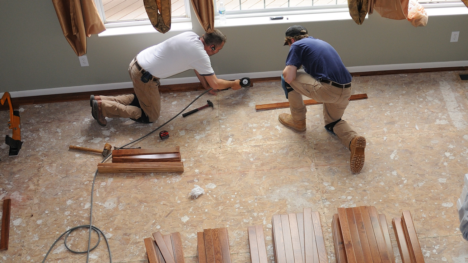 Two men installing hardwood flooring over sub-flooring